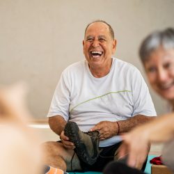 Image of a man in front laughing at the end of a yoga class for older people sitting on the floor on his yoga carpet with his shoe in his hand